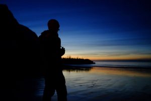 An outline of a person wearing a back pack looking out to sea. It's almost dark, on the horizon is the last of the daylight.