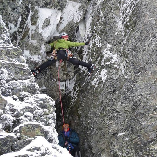 Photo of me and my belayer, I am  bridging my feet and hands across wide crack in the rock. My right leg is twisted so that my foot faces down